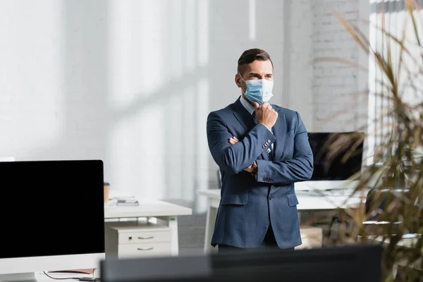 Thoughtful executive in medical mask looking away, while standing in office with blurred plant on foreground — Stock Photo