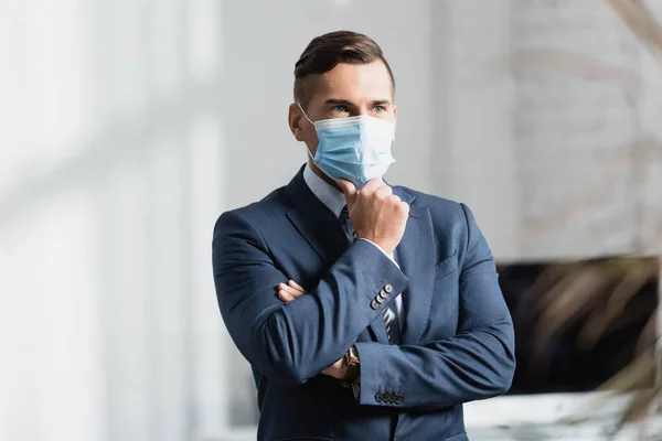 Продуманий бізнесмен в медичній масці, дивлячись вбік, стоячи в офісі на розмитому передньому плані — стокове фото