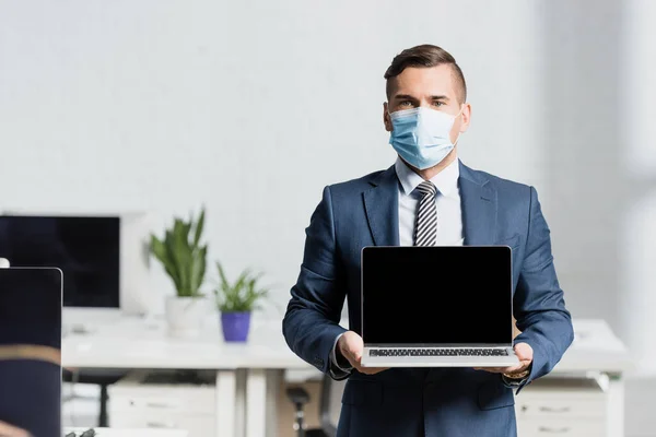 Vue de face de l'homme d'affaires tenant ordinateur portable avec écran blanc avec bureau flou sur fond — Photo de stock