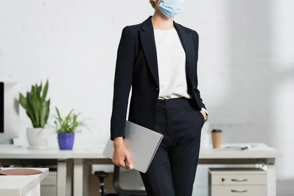 Vista ritagliata di donna d'affari con mano in tasca che tiene il computer portatile, mentre in piedi in ufficio su sfondo sfocato — Foto stock