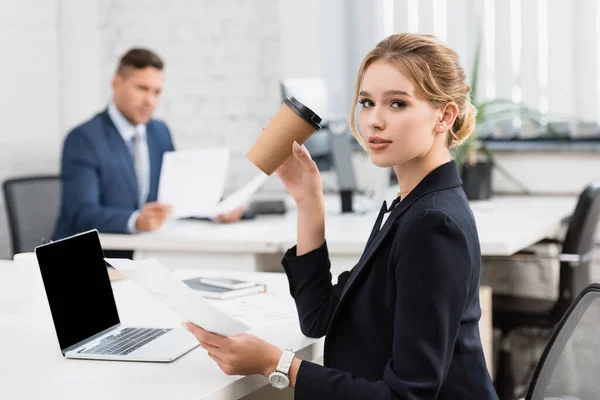 Geschäftsfrau mit Pappbecher blickt in die Kamera, während sie am Arbeitsplatz Papierblätter auf verschwommenem Hintergrund hält — Stockfoto