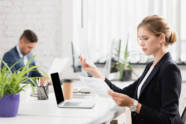 Empresaria enfocada mirando gráficos en hojas de papel, mientras está sentada en el lugar de trabajo sobre fondo borroso - foto de stock