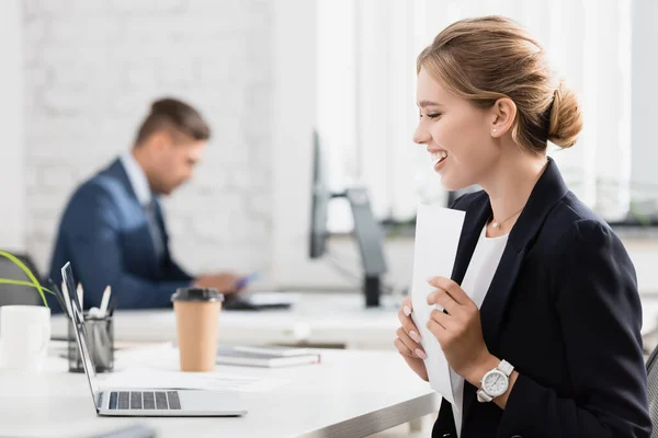 Femme d'affaires heureuse avec feuille de papier regardant ordinateur portable, tout en étant assis sur le lieu de travail sur fond flou — Photo de stock