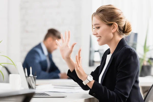 Happy businesswoman with waving hands looking at laptop, while sitting at workplace with blurred colleague on background — Stock Photo