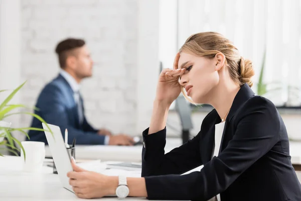 Tired businesswoman with hand near eyes looking at laptop, while sitting at workplace on blurred background — Stock Photo