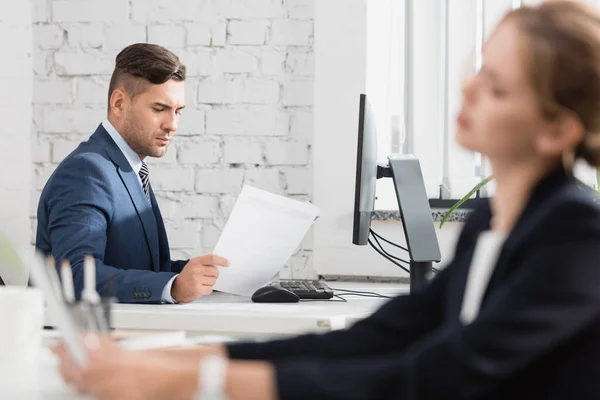 Konzentrierter Geschäftsmann blickt auf Papierbogen, während er am Tisch auf verschwommenem Vordergrund sitzt — Stockfoto