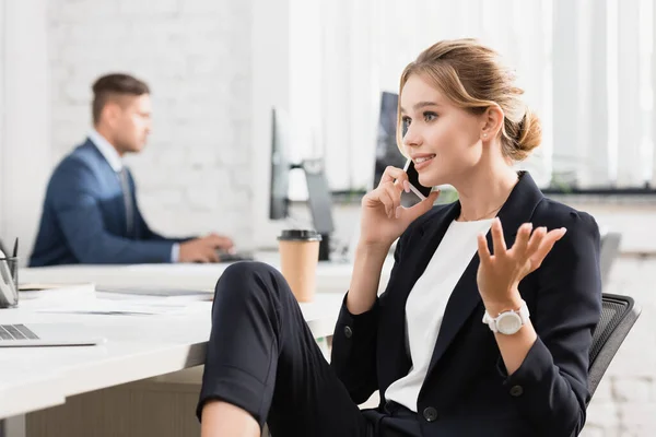 Positive businesswoman gesturing, while talking on mobile phone at workplace on blurred background — Stock Photo