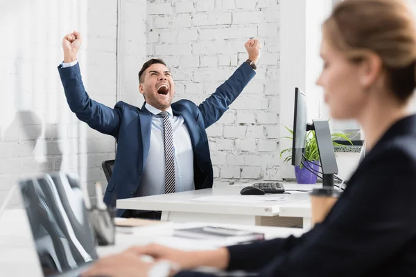 Excited executive with yes gesture sitting at workplace with blurred colleague on foreground — Stock Photo