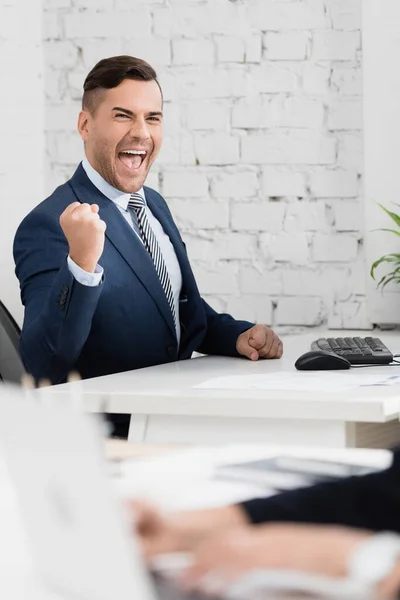 Homme d'affaires excité avec un geste oui regardant la caméra, assis à table au bureau sur le premier plan flou — Photo de stock