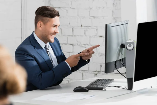 Веселый бизнесмен показывает пальцами, глядя на ноутбук на рабочем месте на размытом переднем плане — стоковое фото