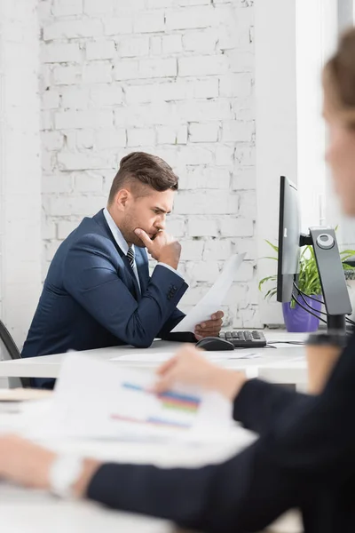 Nachdenklicher Geschäftsmann blickt auf Papierbogen, während er am Arbeitsplatz auf verschwommenem Vordergrund sitzt — Stockfoto