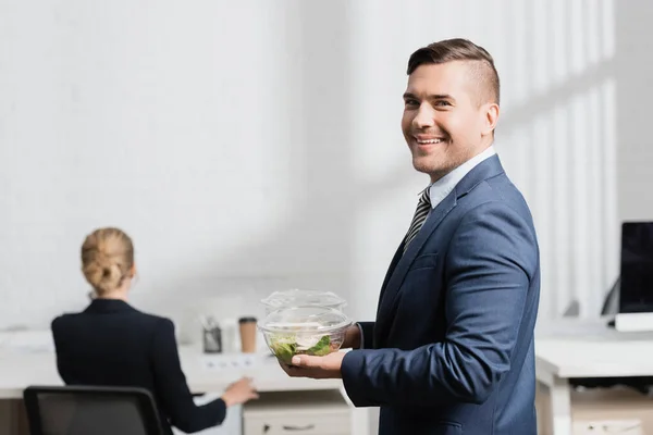 Homme d'affaires souriant avec repas dans des bols en plastique regardant la caméra, tout en restant au bureau sur fond flou — Photo de stock