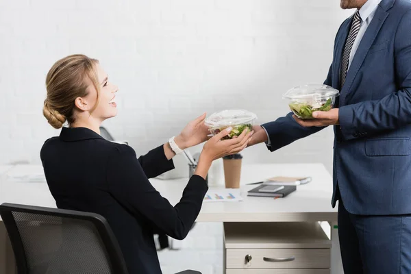 Glückliche Geschäftsfrau nimmt Plastikschüssel mit Mahlzeit vom Kollegen, sitzt am Arbeitsplatz — Stockfoto