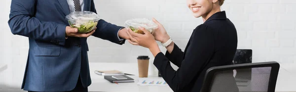 Vue recadrée d'une femme d'affaires souriante prenant un bol en plastique avec de la nourriture de son collègue, assis sur le lieu de travail, bannière — Photo de stock