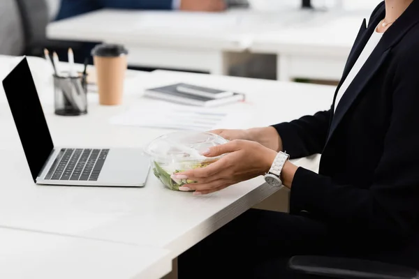 Cropped view of businesswoman holding plastic bowl with meal, while sitting at workplace on blurred background — Stock Photo