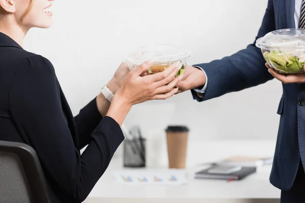 Cropped view of smiling businesswoman taking plastic bowl with meal from co-worker near workplace on blurred background — Stock Photo