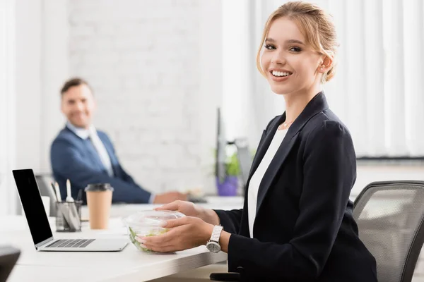 Ejecutivo sonriente con comida en un tazón de plástico mirando a la cámara, mientras está sentado cerca de la computadora portátil en la mesa sobre un fondo borroso - foto de stock