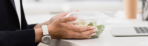 Vue recadrée d'une femme d'affaires tenant un bol en plastique avec de la nourriture au travail sur fond flou, bannière — Photo de stock