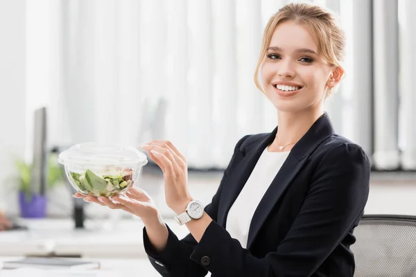 Улыбающаяся деловая женщина с едой в пластиковой миске смотрит в камеру с размытым офисом на заднем плане — стоковое фото