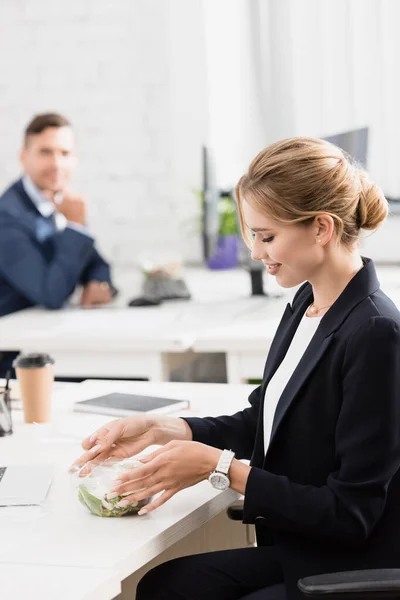 Mujer de negocios sonriente abriendo un tazón de plástico con comida, mientras está sentado en el lugar de trabajo con un colega borroso en el fondo - foto de stock