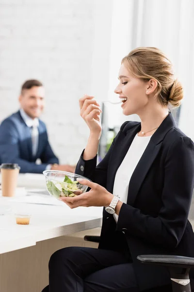 Positive Geschäftsfrau isst Mahlzeit aus Plastikschüssel am Arbeitsplatz in der Pause auf verschwommenem Hintergrund — Stockfoto