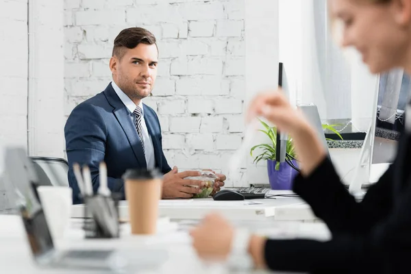 Geschäftsmann mit Mahlzeit in Plastikschüssel blickt in die Kamera, während er am Arbeitsplatz im verschwommenen Vordergrund sitzt — Stockfoto