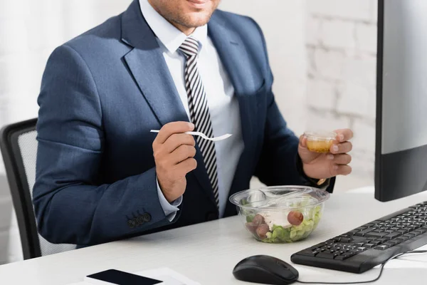 Vista cortada de empresário segurando garfo de plástico e recipiente com molho, enquanto sentado à mesa com refeição em tigela — Fotografia de Stock