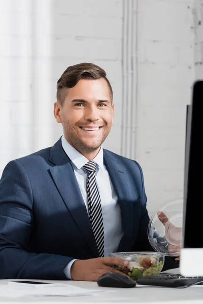Smiling businessman with meal in plastic bowl looking at camera, while sitting at workplace — Stock Photo