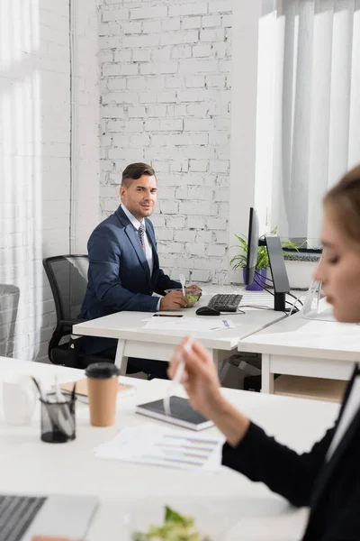 Businessman with meal in plastic bowl and fork looking at colleague, while sitting at workplace on blurred foreground — Stock Photo