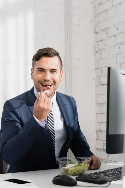 Hombre de negocios sonriente mirando a la cámara, mientras come comida de un tazón de plástico en el lugar de trabajo - foto de stock