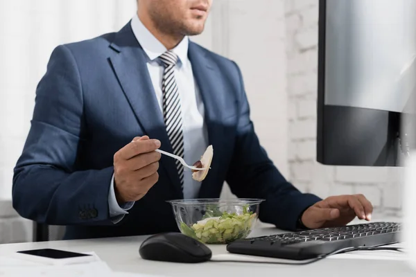 Vista recortada del hombre de negocios comiendo comida de un tazón de plástico, mientras escribe en el teclado de la computadora en el lugar de trabajo - foto de stock