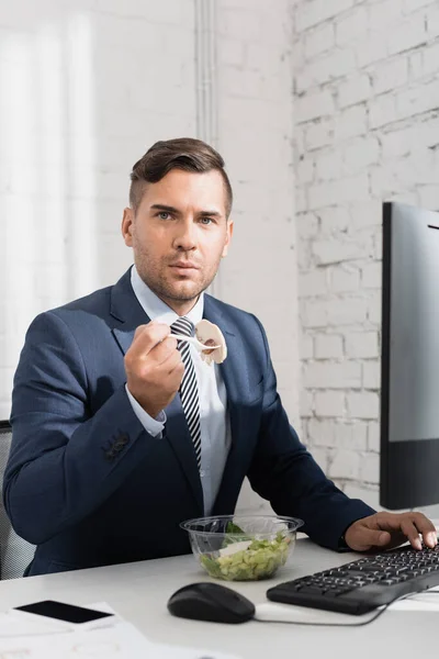 Empresario comiendo comida de un tazón de plástico, mientras mira la cámara en el lugar de trabajo con dispositivos digitales - foto de stock