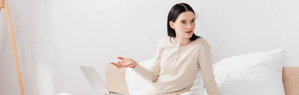 Brünette Frau zeigt mit der Hand in der Nähe von Laptop im Schlafzimmer, Banner — Stockfoto