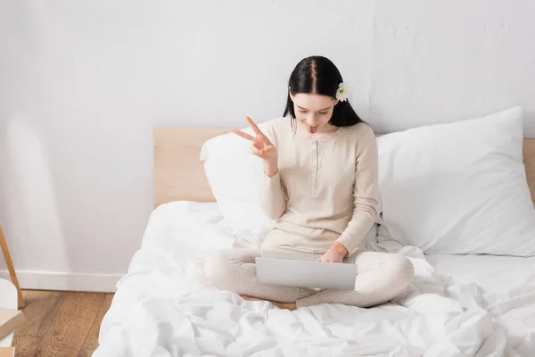 Junge Frau mit Vitiligo und Blume im Haar zeigt Friedenszeichen, während sie Videoanruf auf Laptop im Schlafzimmer — Stockfoto