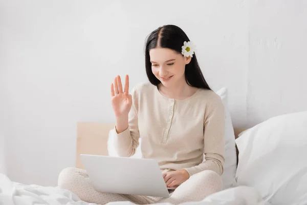 Femme heureuse avec vitiligo et fleur dans les cheveux agitant la main tout en ayant appel vidéo sur ordinateur portable dans la chambre — Photo de stock