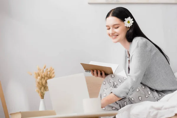 Happy freelancer with vitiligo and flower in hair using laptop and holding notebook near laptop on blurred foreground — Stock Photo