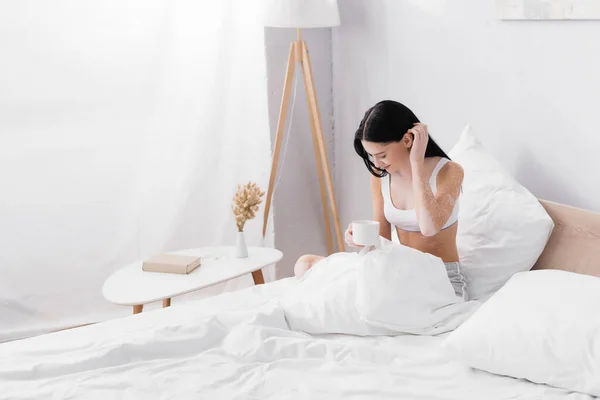 Усміхнена жінка з вітіліго тримає чашку кави, фіксуючи волосся в ліжку — стокове фото