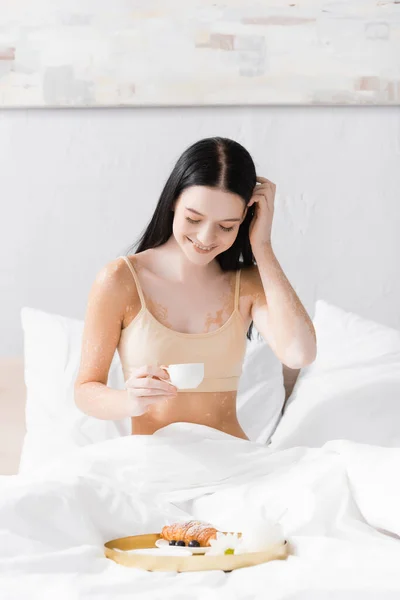 Happy woman with vitiligo holding cup and fixing hair near breakfast tray — Stock Photo