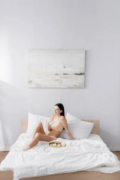 Giovane donna allegra con vitiligine tenendo tazza vicino vassoio colazione sul letto — Foto stock