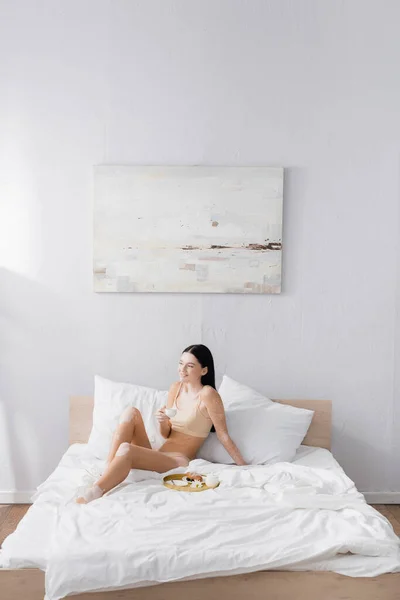 Young cheerful woman with vitiligo holding cup near breakfast tray on bed — Stock Photo