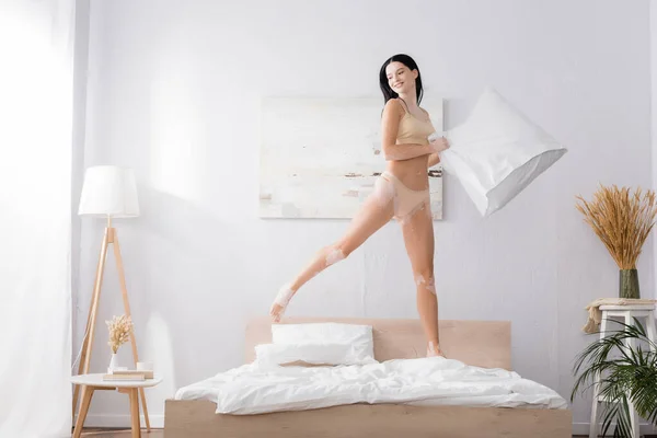 Full length of happy woman with vitiligo jumping with pillow on bed — Stock Photo