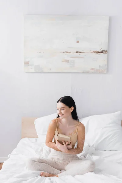 Frau mit Vitiligo hält Buch und sitzt auf Bett im Schlafzimmer — Stockfoto