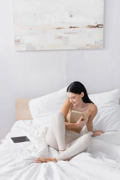Жінка з вітіліго тримає книгу і сидить біля цифрового планшета з порожнім екраном в спальні — стокове фото