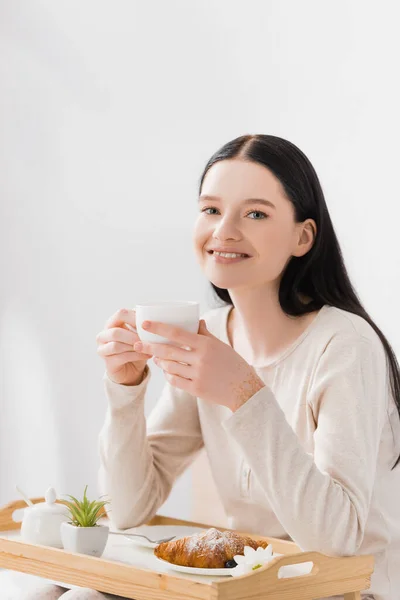 Happy woman with vitiligo holding cup of tea near breakfast on tray — Stock Photo