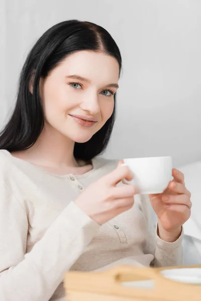 Young happy woman with vitiligo holding cup of coffee near tray on blurred foreground — Stock Photo