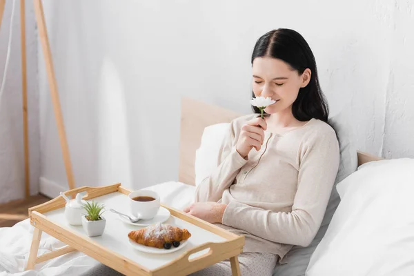Young brunette woman with vitiligo smelling flower near breakfast on tray — Stock Photo