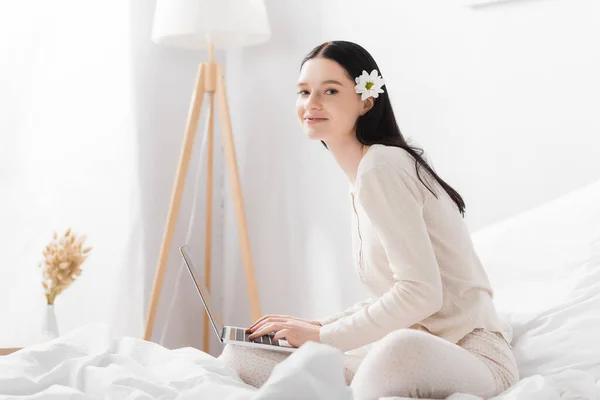 Mulher feliz com vitiligo e flor no cabelo digitando no laptop no quarto — Fotografia de Stock