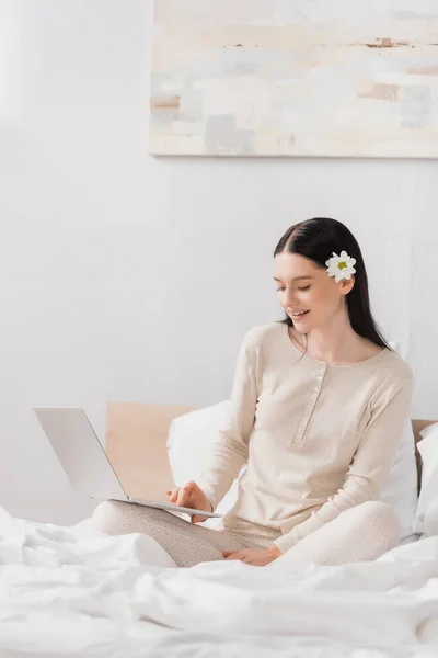 Happy woman with vitiligo and flower in hair using laptop in bedroom — Stock Photo