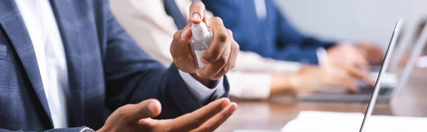 Vista recortada del empresario afroamericano que aplica desinfectante en las manos en el lugar de trabajo sobre fondo borroso, pancarta — Stock Photo