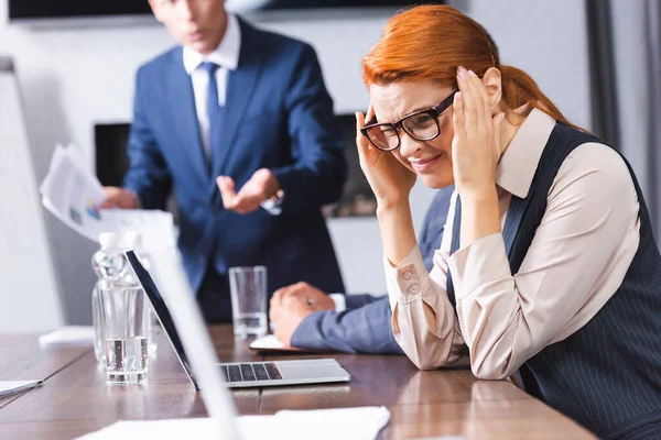 Scared redhead businesswoman with hands near head squinting while sitting near colleagues on blurred background — Stock Photo